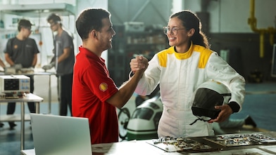 Homem com camisa vermelha da Shell e mulher com macacão branco segurando um capacete apertam as mãos em um laboratório, com equipamentos eletrônicos e outras pessoas trabalhando ao fundo.