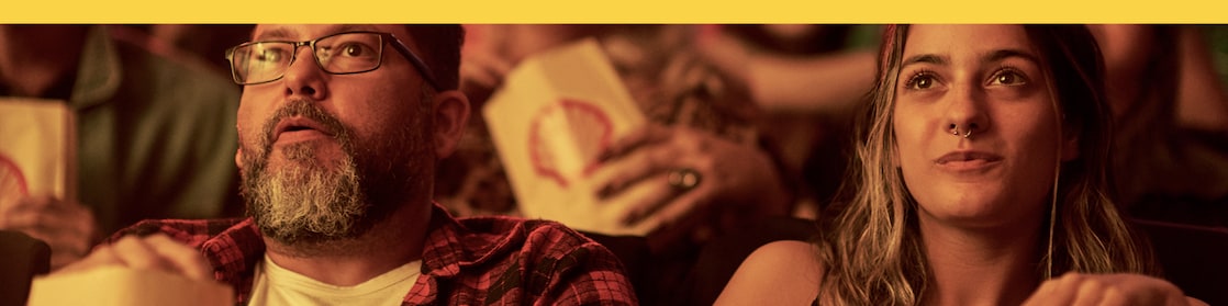 Na foto vemos duas pessoas sentadas em poltronas de cinema, com um saquinho de pipoca com a marca Shell