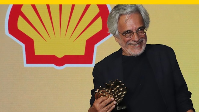 Na foto, vemos Aderbal Freire Filho usando óculos, blazer preto e camisa preta, enquanto segura troféu do Prêmio Shell de Teatro. Ao fundo, vemos em monitor um fundo amarelo com logo da Shell.