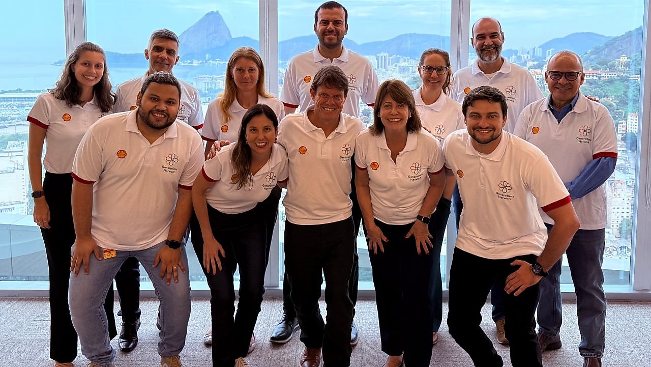 Colaboradores da Shell Brasil posam sorridentes em grupo durante o encontro da rede de afinidade Generations no escritório do Rio de Janeiro. Todos vestem camisetas brancas com o logo da Shell e da rede Generations, com o Pão de Açúcar ao fundo visto pelas janelas panorâmicas.