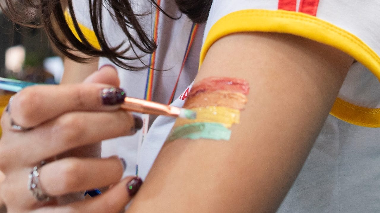Uma pessoa pinta, em seu braço, a bandeira com as cores do arco-íris: vermelho , laranja, amarelo, verde, azul e roxo.