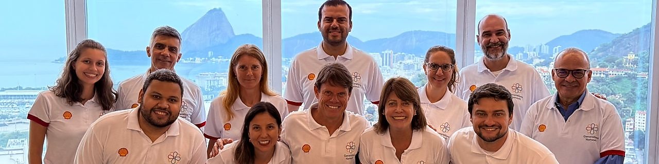 Colaboradores da Shell Brasil posam sorridentes em grupo durante o encontro da rede de afinidade Generations no escritório do Rio de Janeiro. Todos vestem camisetas brancas com o logo da Shell e da rede Generations, com o Pão de Açúcar ao fundo visto pelas janelas panorâmicas.