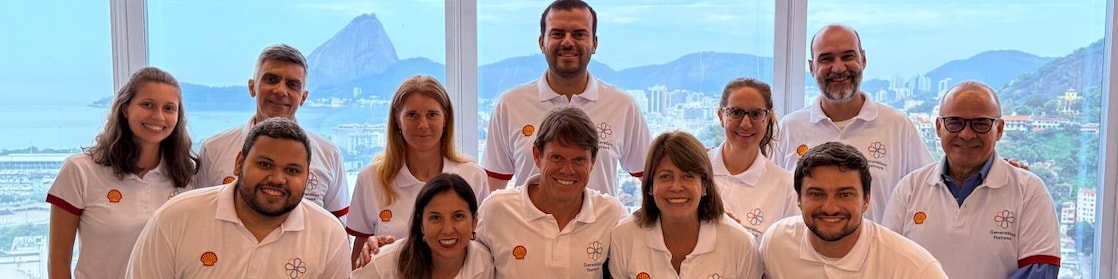 Colaboradores da Shell Brasil posam sorridentes em grupo durante o encontro da rede de afinidade Generations no escritório do Rio de Janeiro. Todos vestem camisetas brancas com o logo da Shell e da rede Generations, com o Pão de Açúcar ao fundo visto pelas janelas panorâmicas.