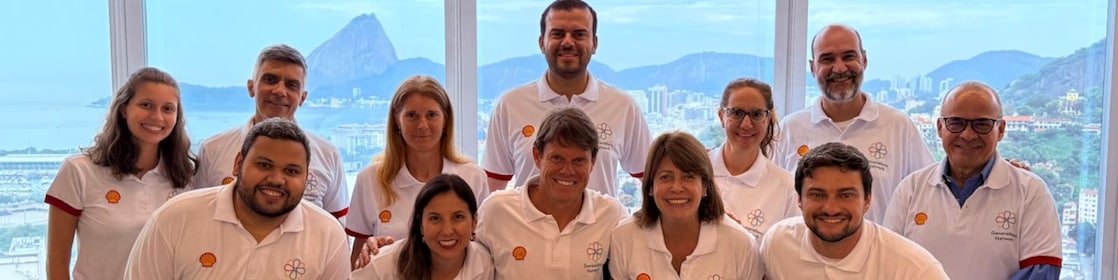 Colaboradores da Shell Brasil posam sorridentes em grupo durante o encontro da rede de afinidade Generations no escritório do Rio de Janeiro. Todos vestem camisetas brancas com o logo da Shell e da rede Generations, com o Pão de Açúcar ao fundo visto pelas janelas panorâmicas.