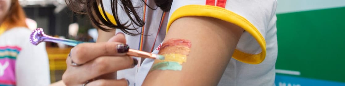 Uma pessoa pinta, em seu braço, a bandeira com as cores do arco-íris: vermelho , laranja, amarelo, verde, azul e roxo.