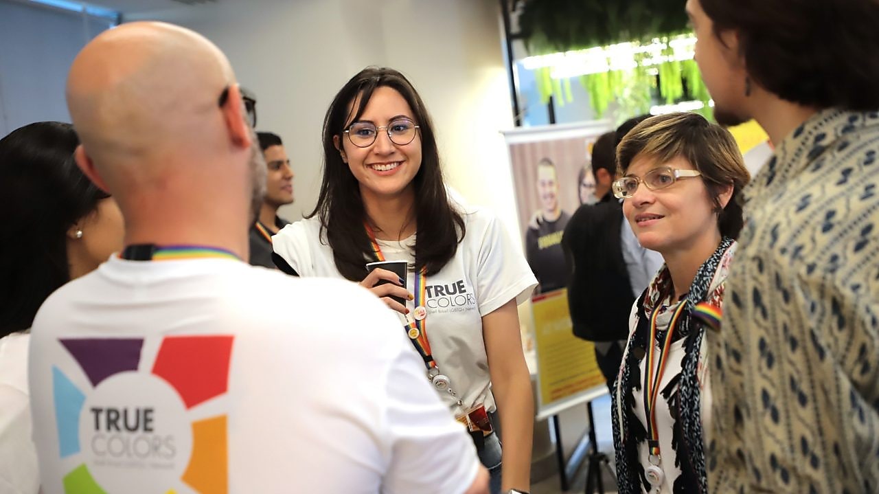 No foco, é possível ver duas mulheres. À esquerda, em primeiro plano, desfocado, é possível ver uma pessoa utilizando uma camisa com a identidade visual da TRUEColors.