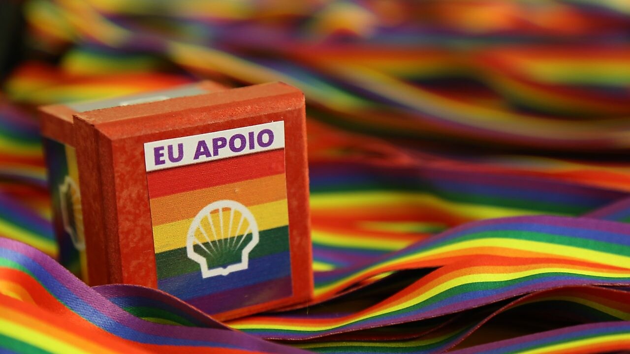 Em uma superfície é possível ver várias cordas com as cores da bandeira do arco-íris: vermelho , laranja, amarelo, verde, azul e roxo. Ao centro, uma caixinha leva também as cores da bandeira do orgulho LGBTQIA+, com o logo da Shell por cima e o texto “Eu apoio”.
