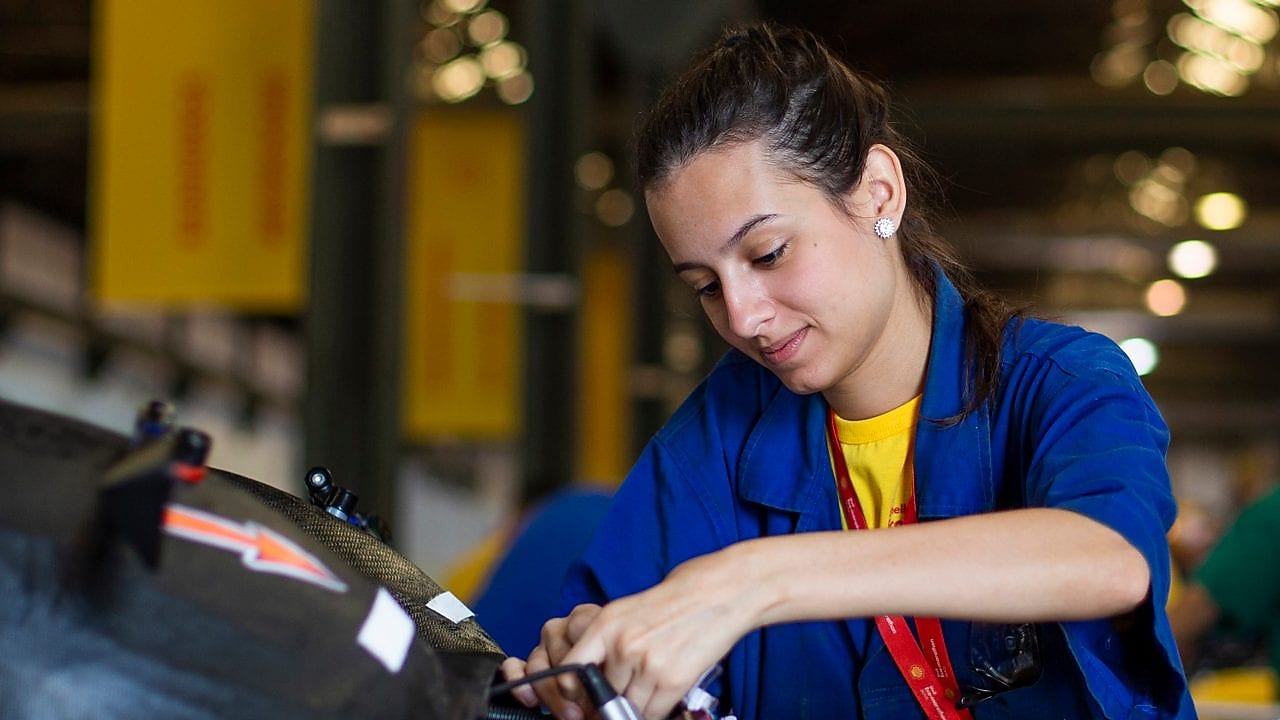 Em um fundo amarelo, vemos três ilustrações de mulheres com uniforme da Shell posicionadas lado a lado. A primeira está com um jaleco vermelho e fones de proteção, a segunda com o uniforme branco e a terceira com uniforme vermelho, fone de proteção e capacete de proteção branco.