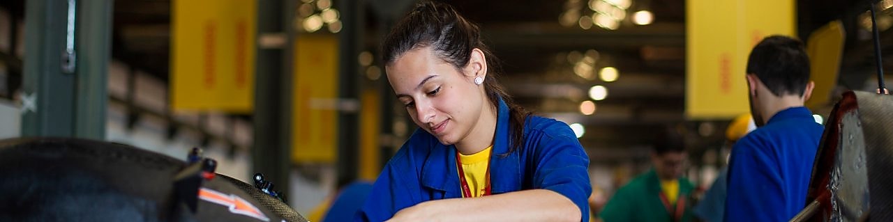 Em um fundo amarelo, vemos três ilustrações de mulheres com uniforme da Shell posicionadas lado a lado. A primeira está com um jaleco vermelho e fones de proteção, a segunda com o uniforme branco e a terceira com uniforme vermelho, fone de proteção e capacete de proteção branco.