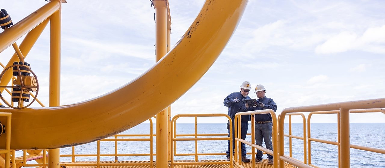 Dois trabalhadores com capacetes e uniformes estão em uma estrutura metálica elevada, em ambiente offshore, sobre o mar. Eles observam ou conferem um equipamento enquanto permanecem apoiados no corrimão da plataforma. A estrutura é pintada de amarelo e o oceano aparece ao fundo. 