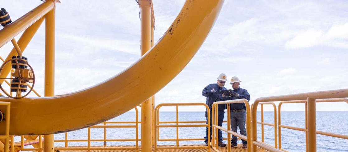 Dois trabalhadores com capacetes e uniformes estão em uma estrutura metálica elevada, em ambiente offshore, sobre o mar. Eles observam ou conferem um equipamento enquanto permanecem apoiados no corrimão da plataforma. A estrutura é pintada de amarelo e o oceano aparece ao fundo.