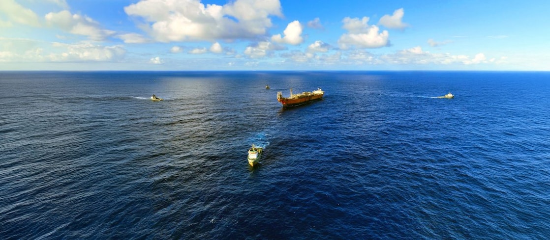 Vista aérea de uma embarcação de grande porte navegando em mar aberto, acompanhada por três embarcações menores ao redor. O oceano azul ocupa a maior parte da imagem, enquanto o horizonte se encontra ao fundo sob um céu claro com nuvens brancas espalhadas. As embarcações deixam rastros na água, destacando o deslocamento em meio à imensidão do mar.