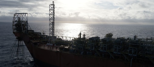 Vista lateral de uma unidade flutuante de produção em alto-mar, com estruturas industriais e equipamentos distribuídos sobre o convés. A embarcação aparece em primeiro plano sobre águas escuras, enquanto o sol reflete na superfície do oceano ao fundo. O céu está parcialmente encoberto por nuvens, criando um contraste entre a luminosidade do horizonte e a silhueta da plataforma.
