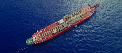 Vista aérea de uma embarcação de grande porte em alto-mar, navegando sobre águas azuis profundas. O navio, de cor vermelha, possui diversas estruturas industriais e tubulações em seu convés, além de uma área de pouso para helicópteros na popa. A superfície do oceano reflete a luz do sol ao redor da embarcação.