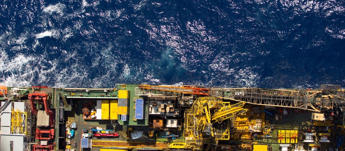 Vista aérea de uma plataforma de petróleo offshore, mostrando estruturas industriais com guindastes, tubulações e equipamentos coloridos em amarelo, vermelho e verde, posicionada sobre o oceano azul com reflexos de luz solar na superfície das ondas.