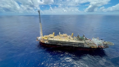 Vista aérea de uma plataforma flutuante de produção de petróleo e gás (FPSO) em alto-mar. A embarcação ocupa a parte inferior da imagem, com um convés repleto de tubulações, módulos de processamento, guindastes e uma torre elevada na extremidade esquerda. À direita, destaca-se um heliponto com a letra "H". Ao redor, o oceano calmo se estende até o horizonte sob um céu azul com nuvens volumosas refletidas na superfície da água.