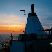 Convés e estrutura superior de um grande navio industrial ao pôr do sol, com chaminé alta, bandeira vermelha hasteada, antenas e equipamentos visíveis, enquanto o mar e o céu alaranjado se estendem ao fundo.