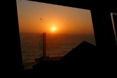 Pôr do sol visto através de uma janela, com o sol baixo no horizonte refletindo tons alaranjados sobre o mar, enquanto as molduras escuras da janela aparecem em silhueta no primeiro plano.