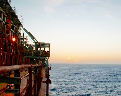 Plataforma de petróleo offshore iluminada ao amanhecer ou entardecer, com estruturas metálicas, tubulações e corrimões à esquerda, enquanto o mar e o céu em tons suaves se estendem até o horizonte à direita.
