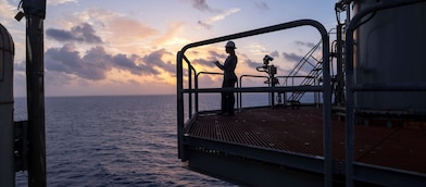 Pessoa usando capacete de segurança consulta um tablet em uma plataforma de petróleo offshore ao amanhecer, com o oceano ao fundo e o céu iluminado por tons alaranjados e azulados. A cena mostra parte da estrutura industrial da plataforma em primeiro plano e o mar no horizonte.