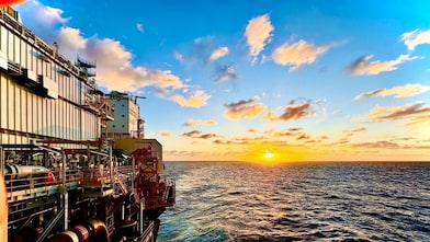 Vista lateral de uma plataforma offshore de petróleo e gás em alto-mar ao pôr do sol. À esquerda, aparecem estruturas metálicas, tubulações e equipamentos industriais; à direita, o oceano se estende até o horizonte, onde o sol baixo ilumina o céu azul com nuvens douradas e reflete sobre a água levemente agitada.