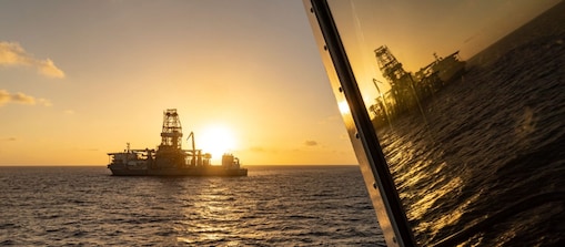 Unidade offshore de perfuração e produção navega sobre o mar ao pôr do sol, com o sol baixo no horizonte iluminando a embarcação. À direita, o reflexo da unidade aparece no vidro de outra embarcação, enquanto a luz dourada se reflete na superfície do oceano.