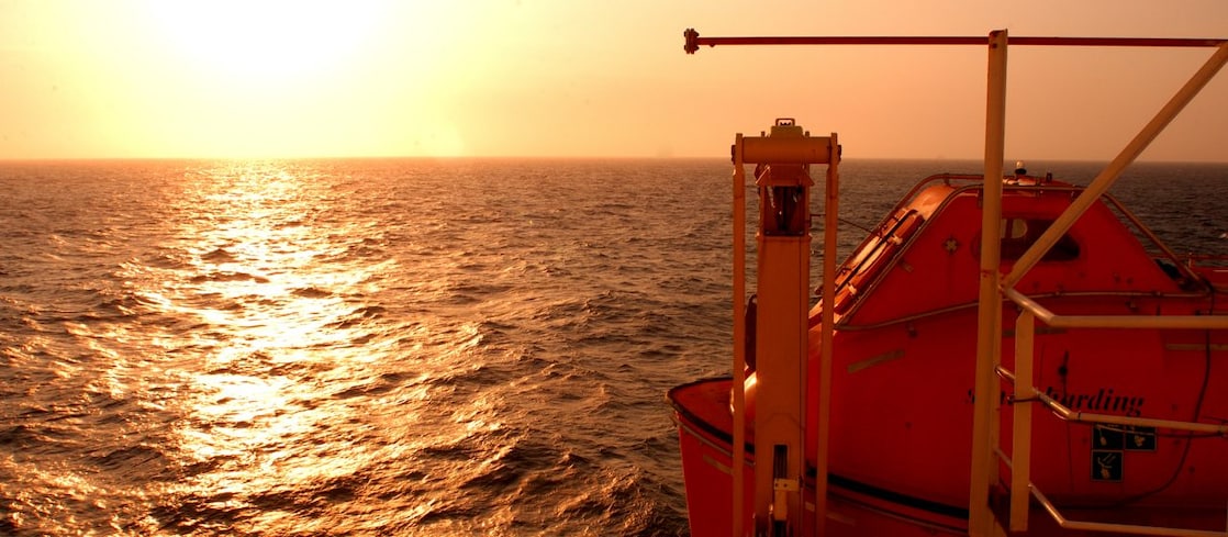 Pôr do sol sobre o mar visto de uma embarcação offshore, com a luz dourada refletindo nas ondas e um bote salva-vidas laranja em primeiro plano.