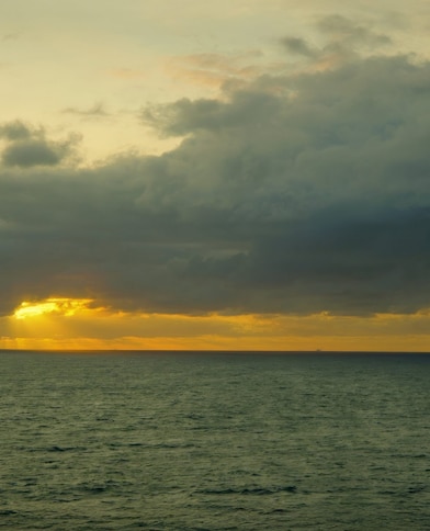 Horizonte marítimo ao entardecer, com o sol parcialmente encoberto por nuvens densas e raios de luz dourada atravessando o céu e iluminando suavemente o mar.