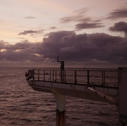 Entardecer no mar com céu em tons suaves de rosa e roxo, nuvens densas no horizonte e parte de uma plataforma offshore projetando-se sobre a água à direita.