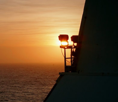 Pôr do sol visto de uma estrutura offshore, com o sol baixo no horizonte iluminando o mar em tons alaranjados, pequenas embarcações ao fundo e parte da plataforma em silhueta à direita.