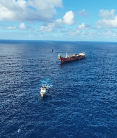 Vista aérea de um navio-tanque no mar aberto, acompanhado por embarcações menores ao redor, sob céu azul com nuvens dispersas.