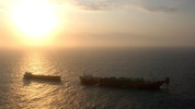 Dois navios no mar ao entardecer, com o sol baixo no horizonte projetando uma luz intensa e dourada sobre a água, criando silhuetas das embarcações.