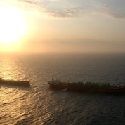 Dois navios no mar ao entardecer, com o sol baixo no horizonte projetando uma luz intensa e dourada sobre a água, criando silhuetas das embarcações.