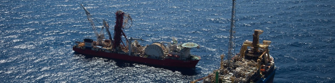 Pôr do sol no mar com o sol baixo no horizonte, céu em tons intensos de laranja e vermelho e parte de uma estrutura industrial offshore em silhueta à esquerda.