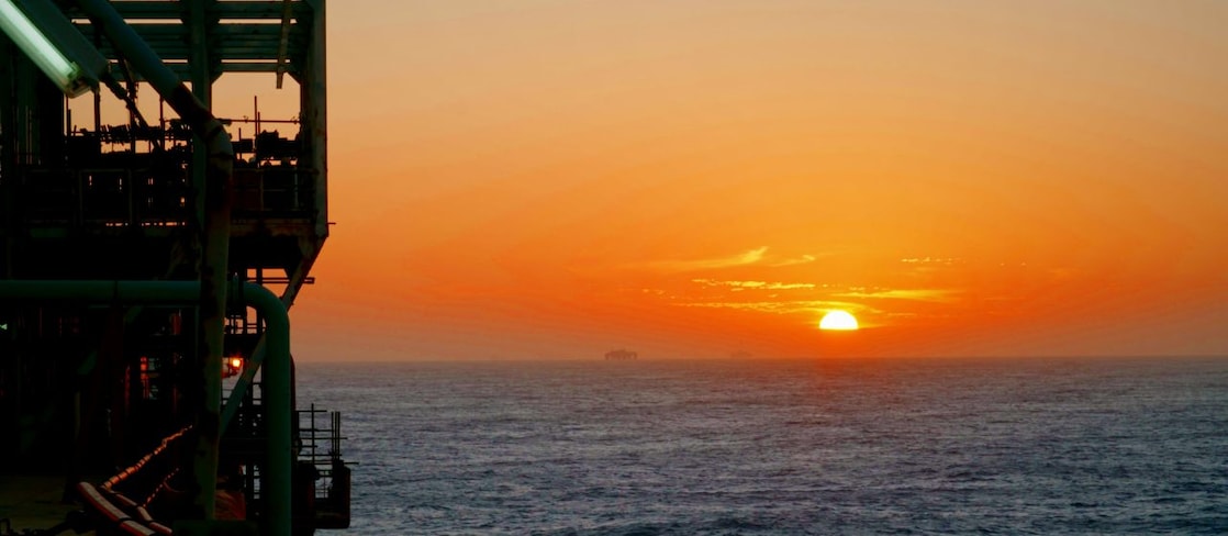 Pôr do sol no mar com o sol baixo no horizonte, céu em tons intensos de laranja e vermelho e parte de uma estrutura industrial offshore em silhueta à esquerda.