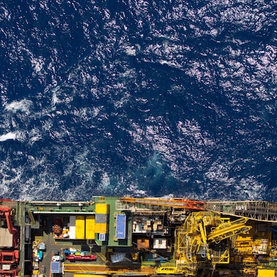 Vista aérea de uma instalação offshore, com estruturas, equipamentos e áreas operacionais na parte inferior da imagem, cercada pelo oceano azul com ondas e reflexos da luz do sol.