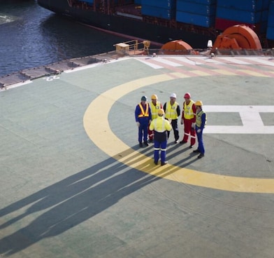 Vista aérea de um heliponto em uma embarcação, onde seis trabalhadores usando capacetes, coletes refletivos e uniformes de segurança estão reunidos. Ao fundo, há um navio de carga e uma pequena embarcação navegando.