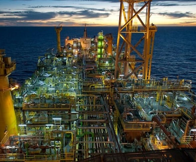 Vista aérea de uma plataforma offshore iluminada ao entardecer, com extensa rede de tubulações, estruturas metálicas e equipamentos industriais, cercada pelo oceano.