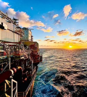 Vista lateral de uma plataforma offshore em alto-mar durante o pôr do sol, com estruturas, tubulações e passarelas industriais à esquerda e o oceano se estendendo até o horizonte sob um céu com nuvens.