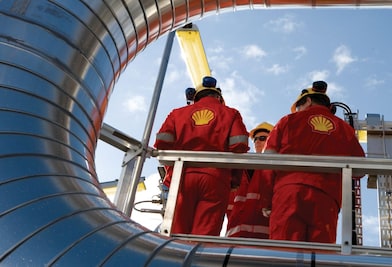 Trabalhadores com uniformes vermelhos, capacetes e equipamentos de proteção atuam em uma instalação industrial, cercados por grandes tubulações metálicas, sob céu azul.