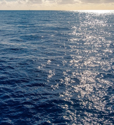 Vista do oceano sob um céu parcialmente nublado, com a luz do sol refletindo sobre a superfície azul da água até o horizonte.