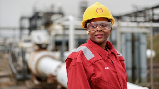 Mulher negra usando capacete amarelo da Shell, óculos de proteção e uniforme vermelho, em ambiente industrial com tubulações e equipamentos ao fundo, olhando de lado.