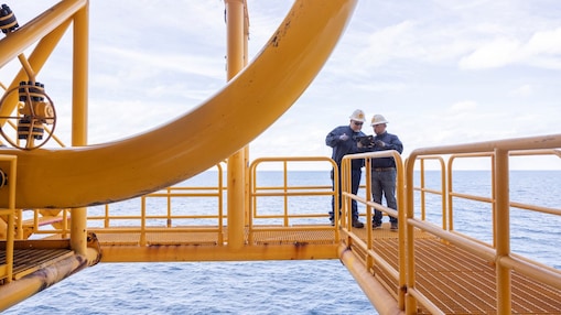 Dois trabalhadores com capacetes e uniformes estão em uma estrutura metálica elevada, em ambiente offshore, sobre o mar. Eles observam ou conferem um equipamento enquanto permanecem apoiados no corrimão da plataforma. A estrutura é pintada de amarelo e o oceano aparece ao fundo.