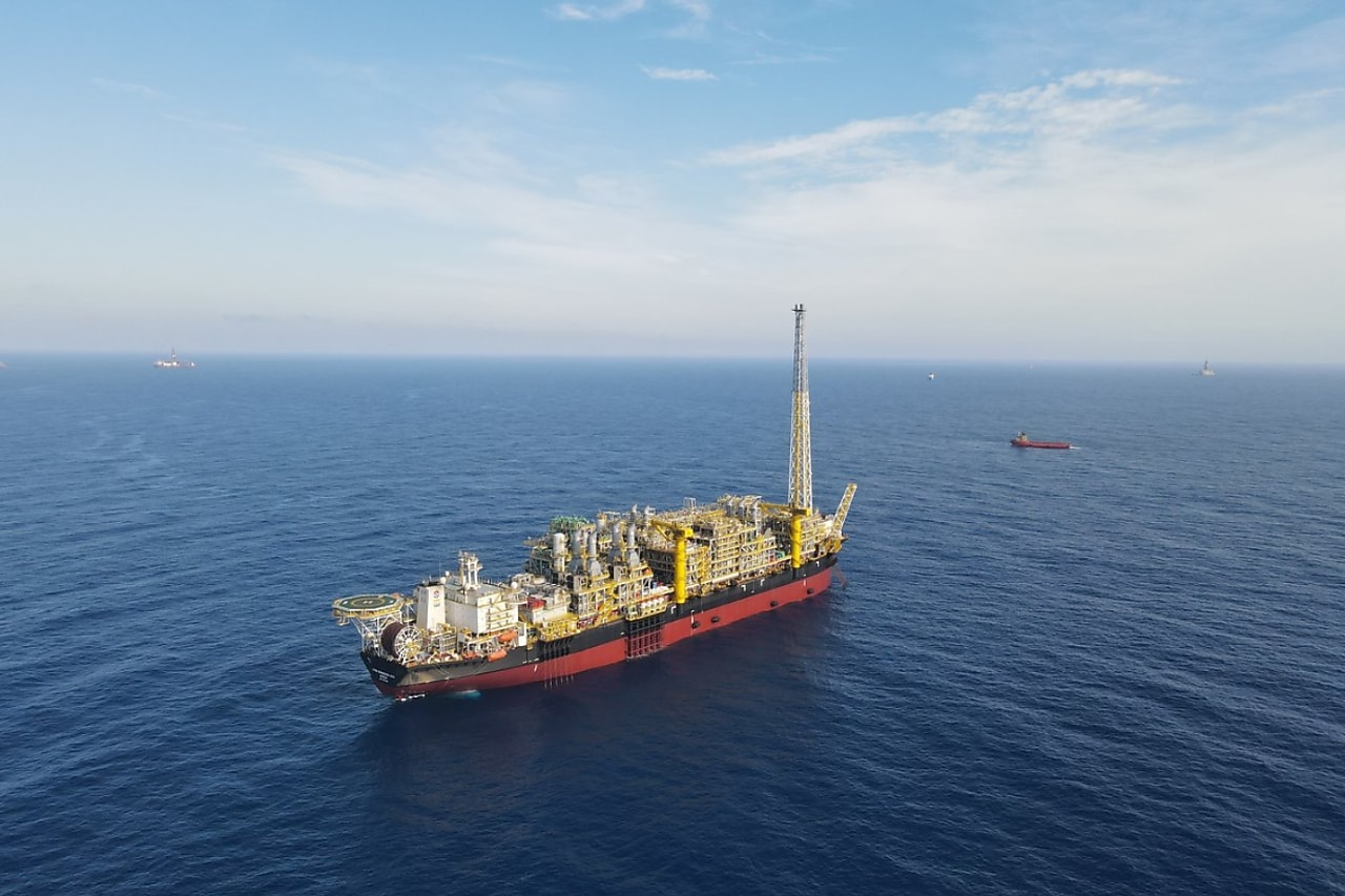 Plataforma flutuante de produção de petróleo (FPSO) em alto-mar, cercada por águas azuis sob um céu parcialmente nublado. A embarcação possui estruturas metálicas complexas e uma torre alta, com outros navios visíveis ao fundo no horizonte.