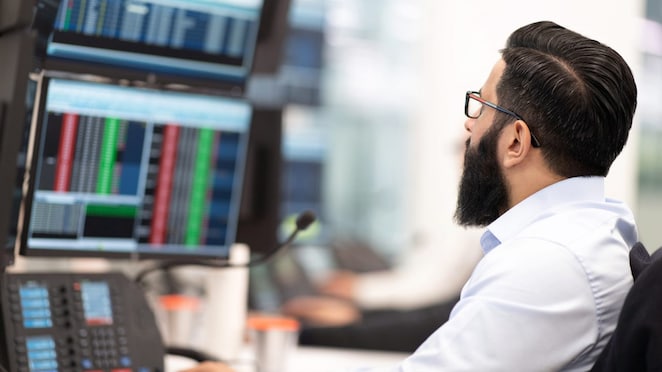 Homem com barba e óculos, vestido com camisa social clara, sentado em frente a várias telas de computador exibindo gráficos e dados financeiros em um ambiente corporativo moderno, provavelmente uma sala de controle ou operação de mercado.