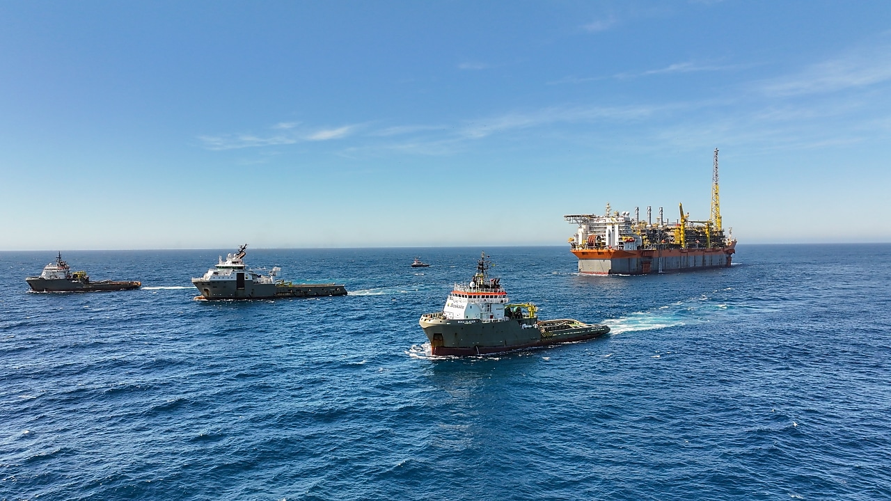 Plataforma de petróleo offshore ao fundo, rodeada por diversos navios de apoio marítimo navegando em águas profundas sob céu azul claro.