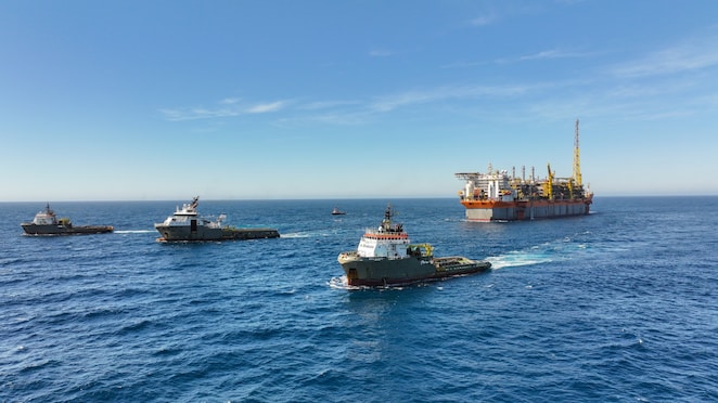 Plataforma de petróleo offshore ao fundo, rodeada por diversos navios de apoio marítimo navegando em águas profundas sob céu azul claro.