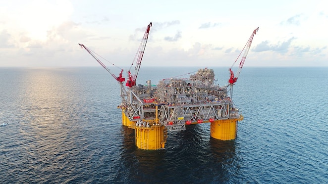 Plataforma de petróleo offshore em operação no mar, com estrutura metálica robusta sustentada por colunas amarelas flutuantes. Três guindastes vermelhos estão posicionados sobre a plataforma. Ao fundo, o céu apresenta nuvens suaves com o sol baixo no horizonte, refletindo na superfície azul do oceano.