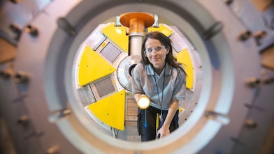 Uma mulher, usando óculos de proteção, está olhando através de uma grande peça circular de equipamento industrial, segurando uma lanterna. Ao fundo, há partes de uma máquina com detalhes amarelos e cinza. A imagem parece ter sido capturada de dentro da estrutura circular, com foco na mulher realizando uma inspeção.