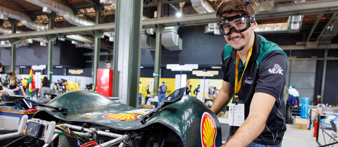 Participante da Shell Eco-marathon trabalha em um veículo de eficiência energética verde, usando óculos e luvas de proteção. O carro está parcialmente aberto, deixando componentes e estruturas internas à mostra, em uma área de boxes do evento.